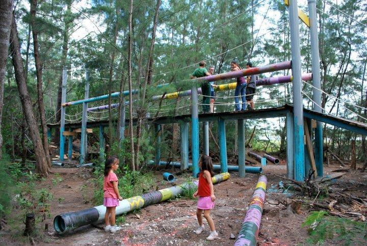 Two young girls in pink dresses are playing in a forested area, while adults are observed on a colorful overhead structure made of pipes and ropes. The background features trees, and colorful painted pipes are scattered on the ground, creating a playful and adventurous scene. Amelia Earhart Park mountain bike trail.