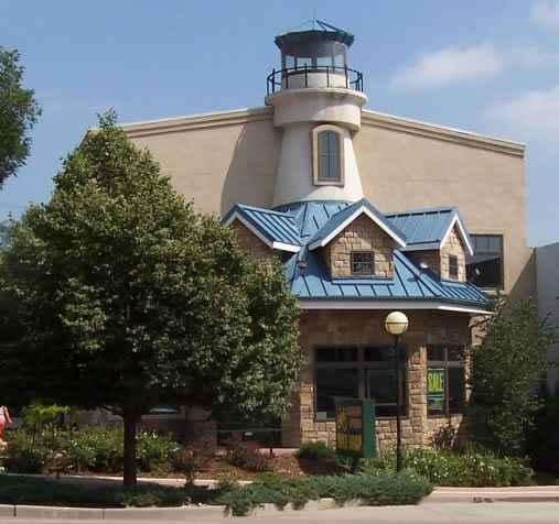 A charming building designed to resemble a lighthouse, featuring a stone facade and a blue roof. The structure is surrounded by well-maintained landscaping with trees and flowers, under a clear blue sky.