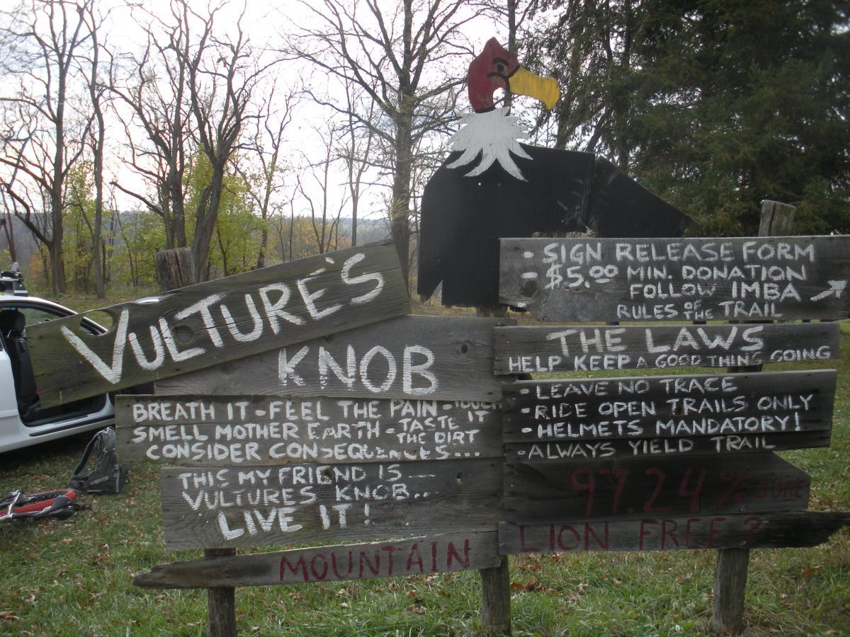 A wooden sign for Vulture's Knob, featuring bold text with trail rules and information. The sign includes a cartoon vulture at the top and provides instructions for visitors, including the need to sign a release form, a minimum donation of $5, and guidelines such as "leave no trace" and "helmets mandatory." The background shows a wooded area and a glimpse of a vehicle. Vultures Knob mountain bike trail.