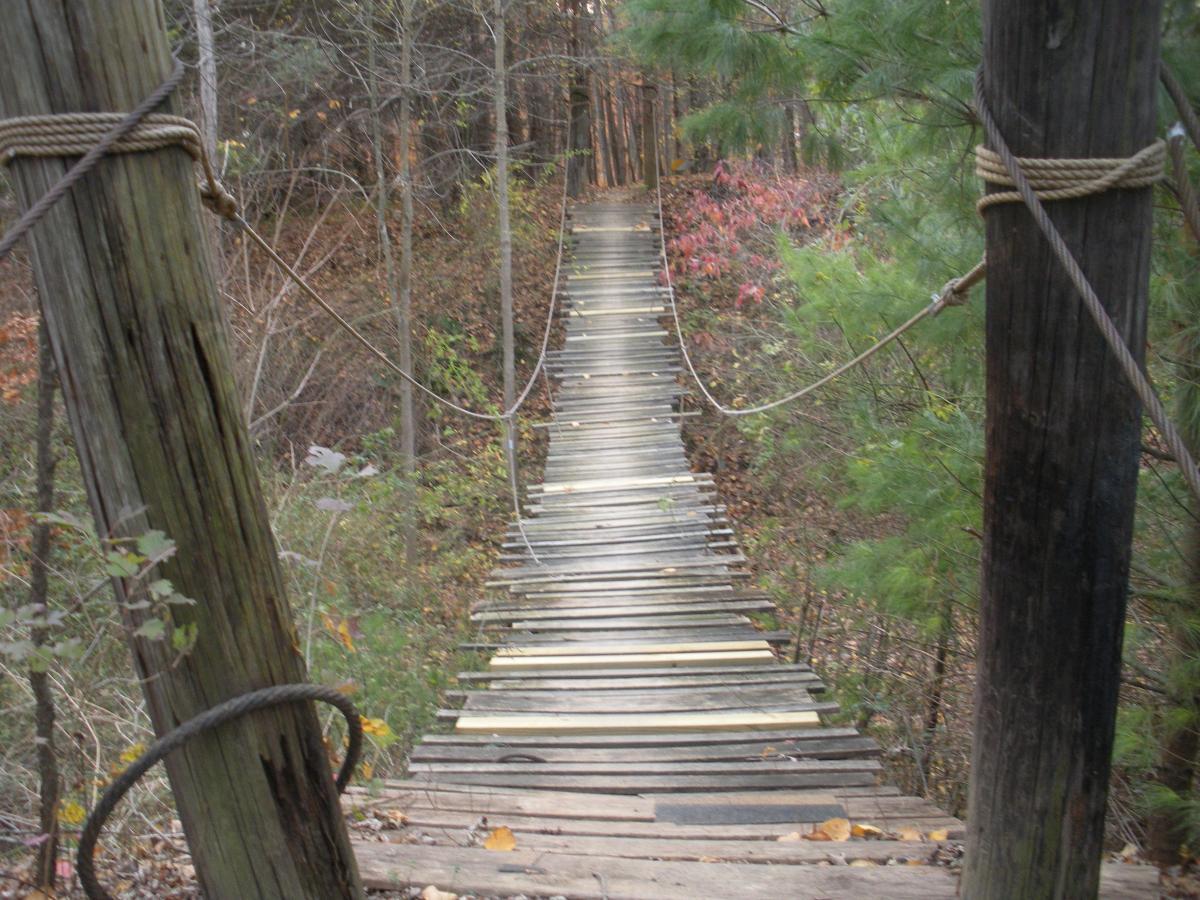 A narrow, wooden suspension bridge stretches over a forested area, lined with trees in various autumn colors. The bridge is supported by sturdy wooden posts and wrapped ropes, providing a rustic and adventurous feel. The view looks down the length of the bridge, emphasizing its height above the ground. The surrounding foliage features hints of red and green leaves, indicating the season. Vultures Knob mountain bike trail.