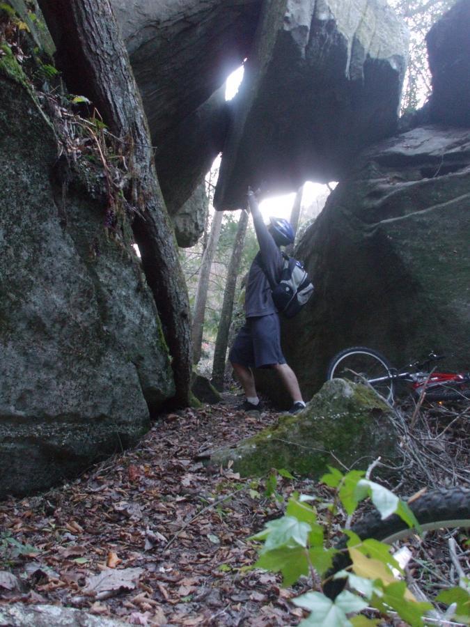 A person in a gray outfit and helmet stands between two large boulders in a forested area, raising one arm towards an overhead rock formation. A bicycle is partially visible on the ground nearby, surrounded by fallen leaves and greenery. Laural Lake To Cumberland Falls (sheltowee Trace) mountain bike trail.