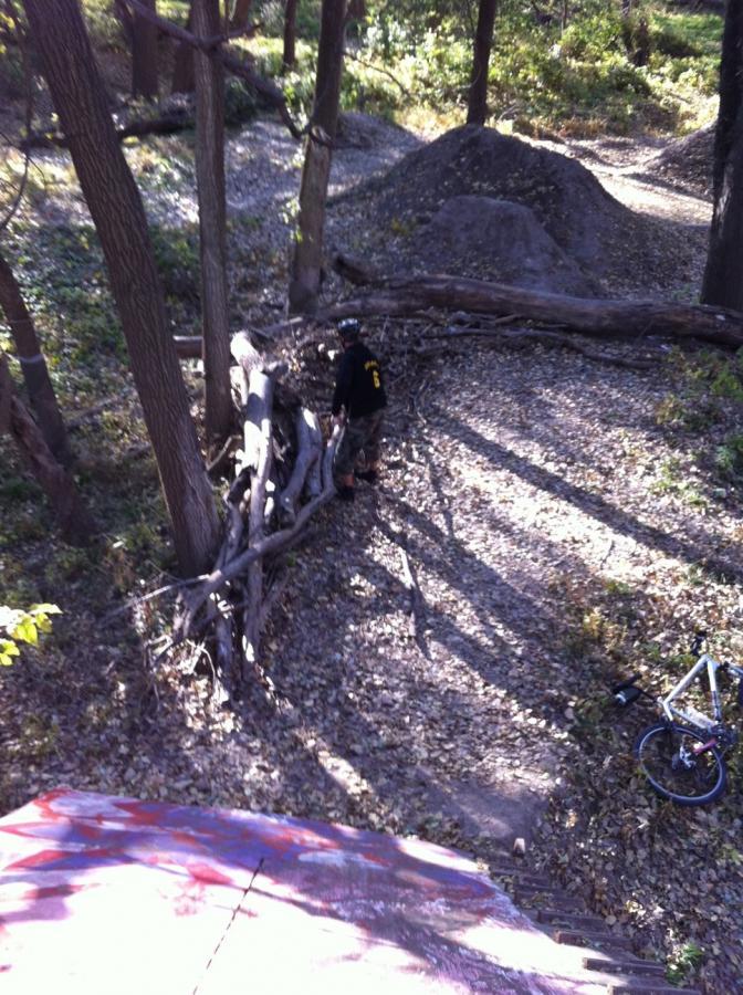 A person wearing a helmet and a black jacket stands next to a pile of logs in a wooded area with fallen leaves. In the background, there is a dirt mound and a mountain bike is resting on the ground nearby. Sunlight filters through the trees, creating patches of light and shadow on the forest floor. Manhattan Pump Track mountain bike trail.
