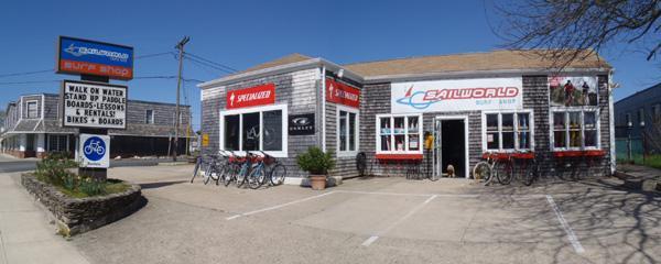 A panoramic view of a storefront featuring "Sailworld" on one side, showcasing various outdoor equipment and bicycles for rent, alongside a sign promoting water activities. The building has a gray wooden exterior and is surrounded by a parking area.