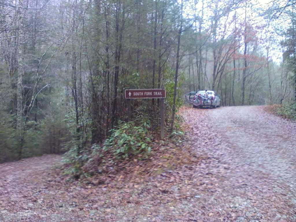 Sign for South Fork Trail at a gravel road intersection, surrounded by dense trees and fallen leaves. A vehicle with bicycles attached can be seen parked near the sign. The scene is slightly misty, creating a serene, natural atmosphere. South Fork Trail mountain bike trail.