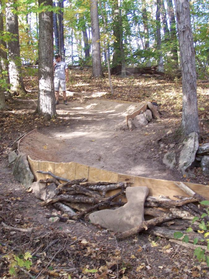 A dirt path winding through a wooded area, surrounded by trees with green and golden leaves. In the foreground, there is a small structure made of wood and branches, possibly a part of a trail or obstacle. A person stands on the path in the background, appearing to explore the area. Rotary Park mountain bike trail.