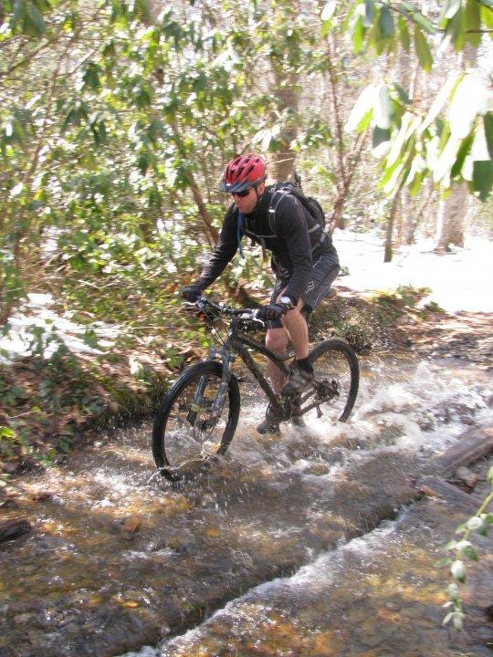 A mountain biker riding through a shallow stream on a wooded trail, splashing water as he navigates the terrain. He is wearing a red helmet and sunglasses, with a backpack on his back, surrounded by lush greenery. Bent Creek mountain bike trail.