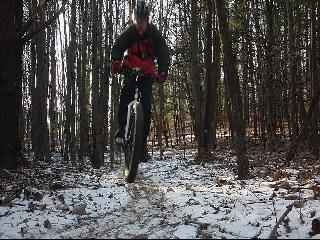 A cyclist riding a mountain bike along a snowy trail in a dense forest, with tall trees lining both sides of the path. The rider is wearing a dark jacket and a helmet, and snow covers parts of the ground, contrasting with the earthy tones of the trail. Moon Lake Park mountain bike trail.