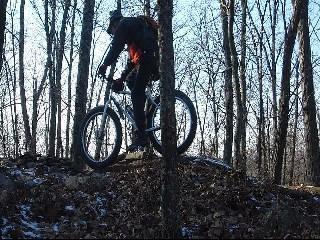 A mountain biker navigating a rocky trail in a wooded area, surrounded by bare trees. The rider is mid-action, with one wheel elevated as they ascend a small hill. The scene captures the essence of outdoor adventure and cycling in nature. Moon Lake Park mountain bike trail.
