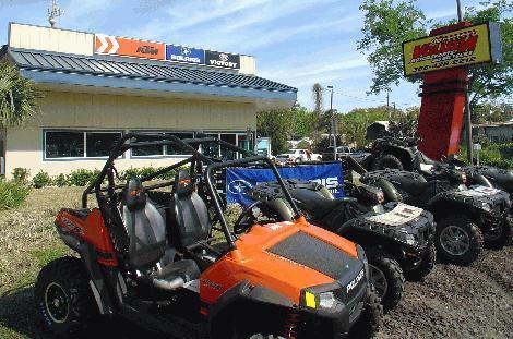 An outdoor view of a vehicle dealership featuring several all-terrain vehicles (ATVs) and a side-by-side UTV parked in front. The dealership building displays a sign with the brand names and logos. There is a banner in the foreground promoting the dealership. The sky is clear and blue, with greenery surrounding the area.