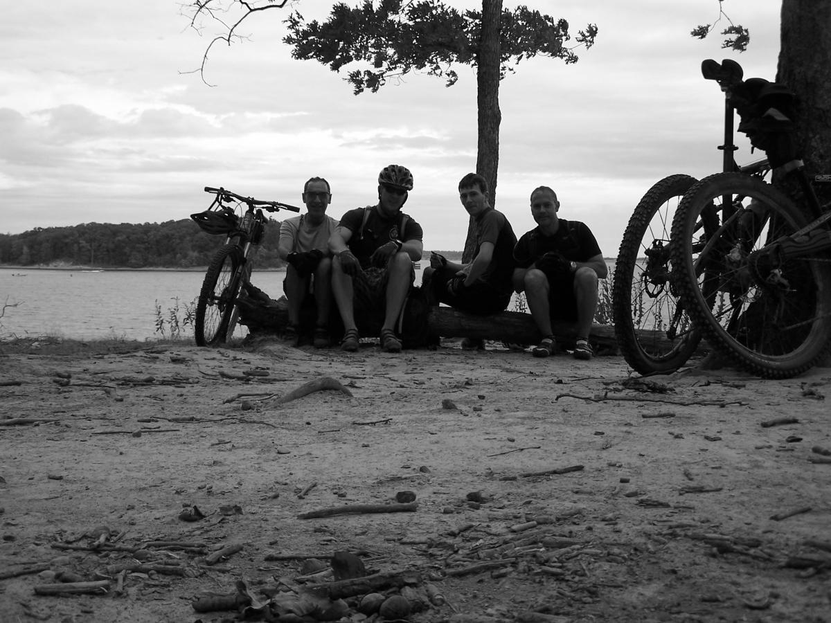 Four individuals sitting on a log by the water, with mountain bikes resting nearby. The image is in black and white, featuring a natural landscape with trees and a cloudy sky in the background. Canal Loop mountain bike trail.