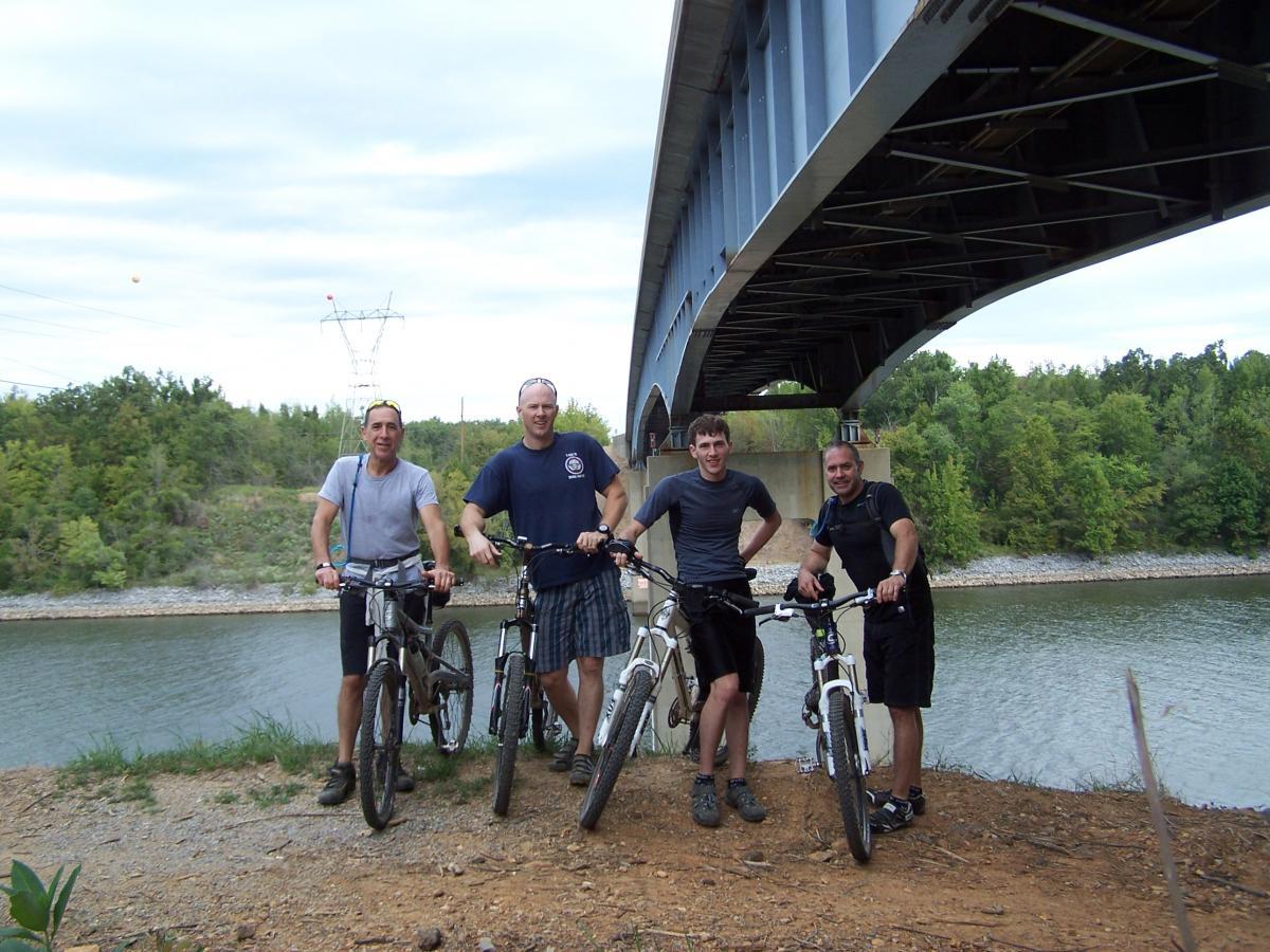 Four men standing with mountain bikes on a gravel bank by a river. They are positioned under a large bridge with greenery in the background and power lines visible in the distance. The sky is partly cloudy, creating a relaxed outdoor atmosphere. Canal Loop mountain bike trail.