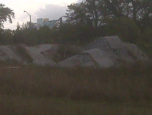 A view of a grassy area featuring mounds of sand or gravel, with a faint outline of a billboard or structure in the background. The image is taken during a cloudy day, adding a muted tone to the scene. Quiet Waters Park mountain bike trail.