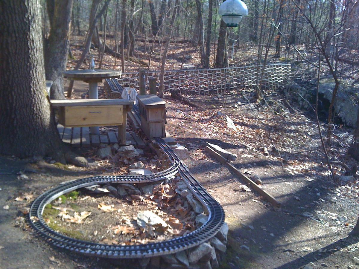 A wooden bench and table are situated near a winding miniature train track in a wooded area with scattered leaves. A vintage-style lamp post can be seen in the background, adding to the tranquil outdoor setting. Needham Town Forest mountain bike trail.