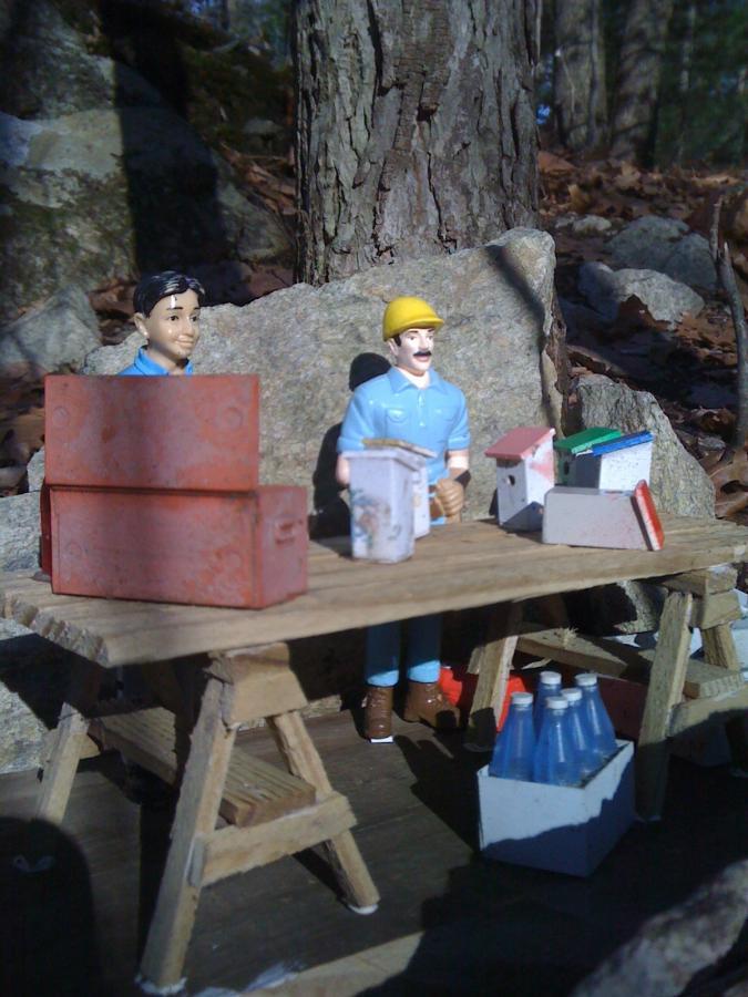 Two toy figures are set up at a rustic wooden table outdoors, surrounded by rocks and leaves. One figure, wearing a blue shirt, is seated while the other, wearing a yellow hard hat and blue shirt, stands beside a display of miniature houses and various small containers, including boxes and bottles. The setting appears to be playful and imaginative, with a focus on crafting or building activities. Needham Town Forest mountain bike trail.