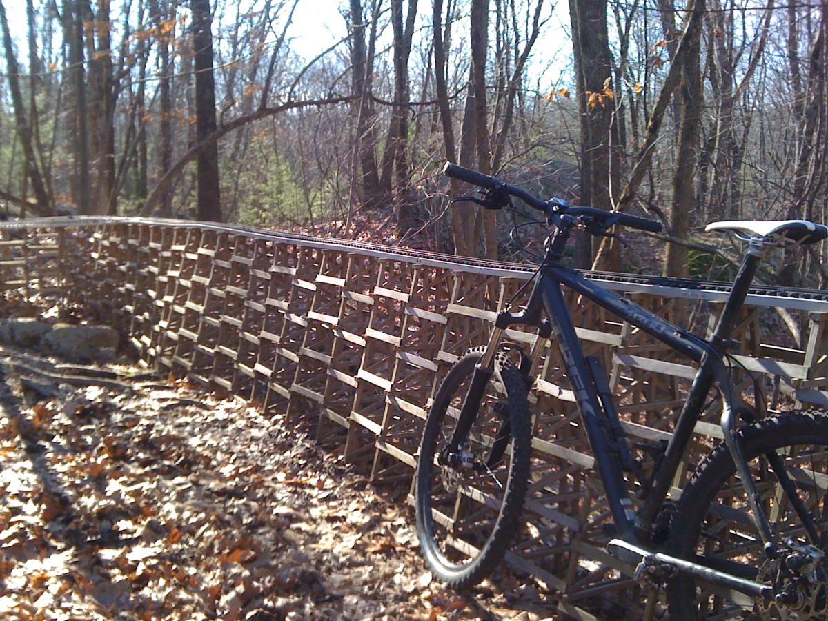 A mountain bike is positioned beside a wooden elevated trail in a forested area, surrounded by trees and fallen leaves on the ground. The trail features a unique lattice design, emphasizing outdoor adventure and biking. Needham Town Forest mountain bike trail.