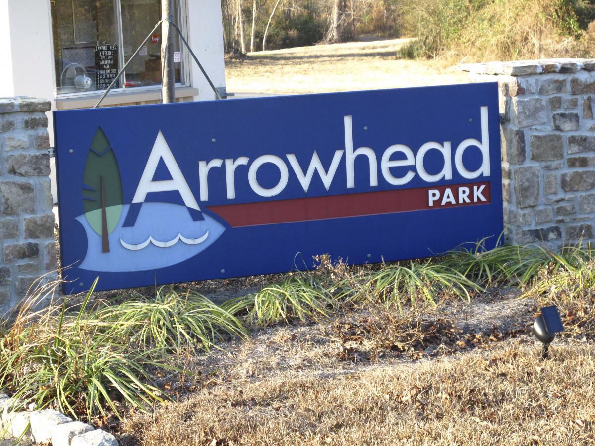 Sign for Arrowhead Park, featuring the name "Arrowhead" in large letters with a stylized design of a fish and a tree. The sign is surrounded by stonework and greenery, indicating the park's entrance. Arrowhead Park mountain bike trail.