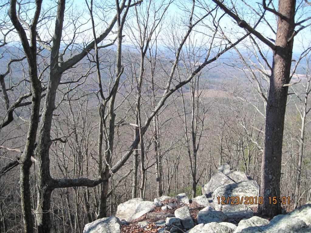 A scenic view from a rocky outcrop overlooking a valley, surrounded by bare trees on a clear day. The landscape features distant hills and open fields, capturing a tranquil, natural setting in winter. Pine Log WMA mountain bike trail.