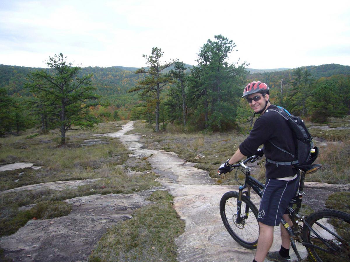 A smiling cyclist wearing a helmet and sunglasses stands next to a mountain bike on a rocky trail surrounded by trees and rolling hills in the background. The landscape features a mix of greenery and autumn foliage. DuPont State Recreational Forest mountain bike trail.
