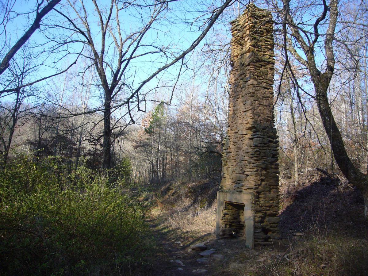 A weathered stone chimney standing alone among trees in a wooded area, with clear blue skies above and a path leading into the forest. Tsali Left Loop mountain bike trail.