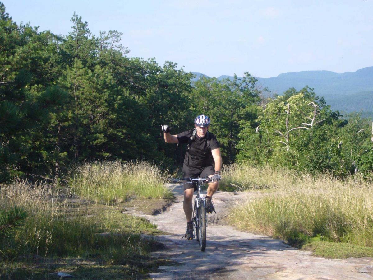 A person riding a mountain bike along a rocky trail in a natural outdoor setting, surrounded by greenery and trees, with mountains visible in the background. The cyclist is wearing a helmet and raising one arm, appearing to express excitement or joy. DuPont State Forest mountain bike trail.