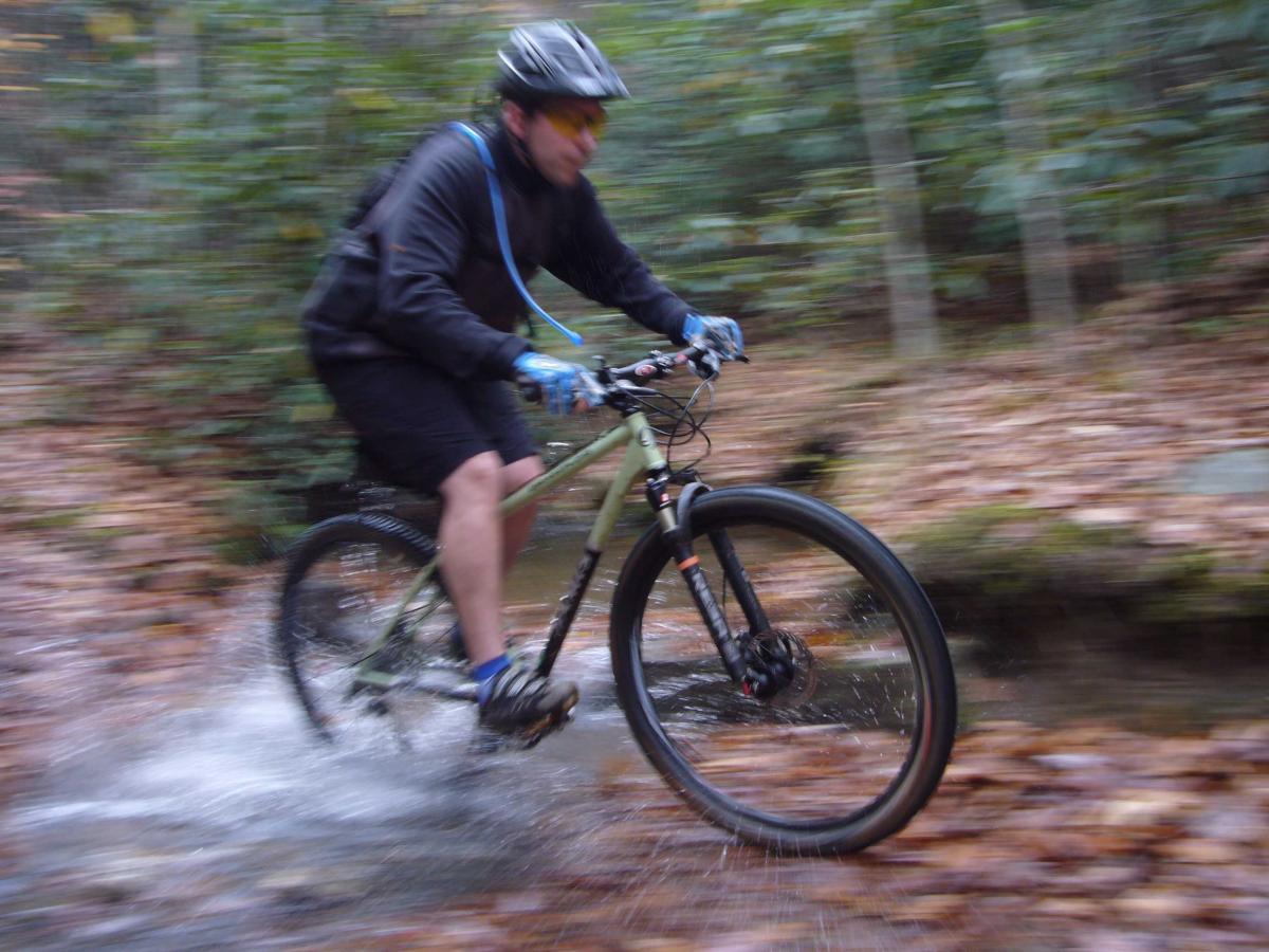 A mountain biker rides through a wooded trail splashing through a shallow stream, surrounded by fallen leaves and blurred greenery, capturing a sense of speed and adventure. Bent Creek mountain bike trail.