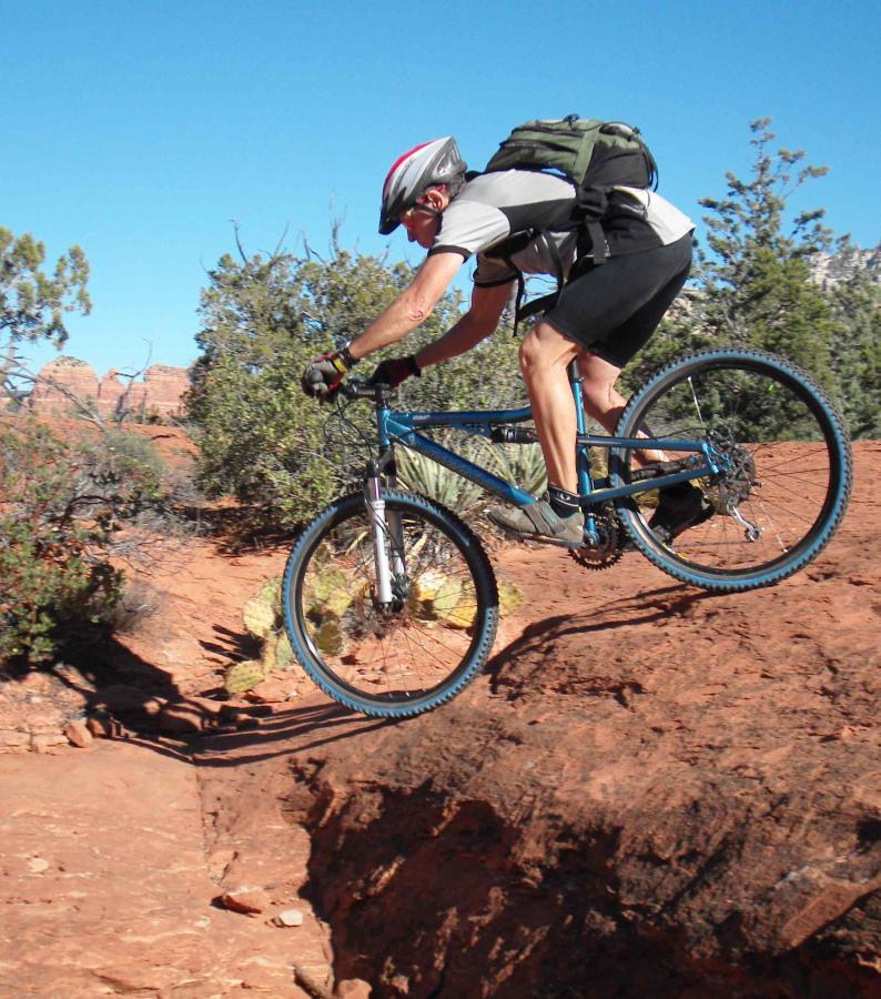 A mountain biker in a helmet and athletic gear jumps over a rocky gap on a dirt trail surrounded by desert vegetation and blue sky. Soldier Wash Trails mountain bike trail.