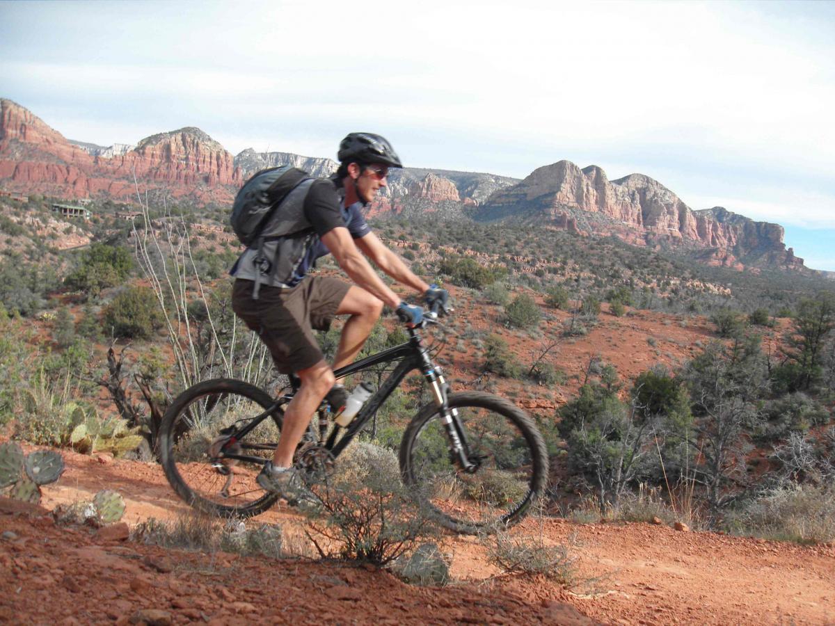 A mountain biker riding along a dirt trail in a rugged desert landscape, with red rock formations and sparse vegetation in the background. The biker is wearing a helmet, a backpack, and casual cycling attire, showcasing an active outdoor experience. Templeton mountain bike trail.