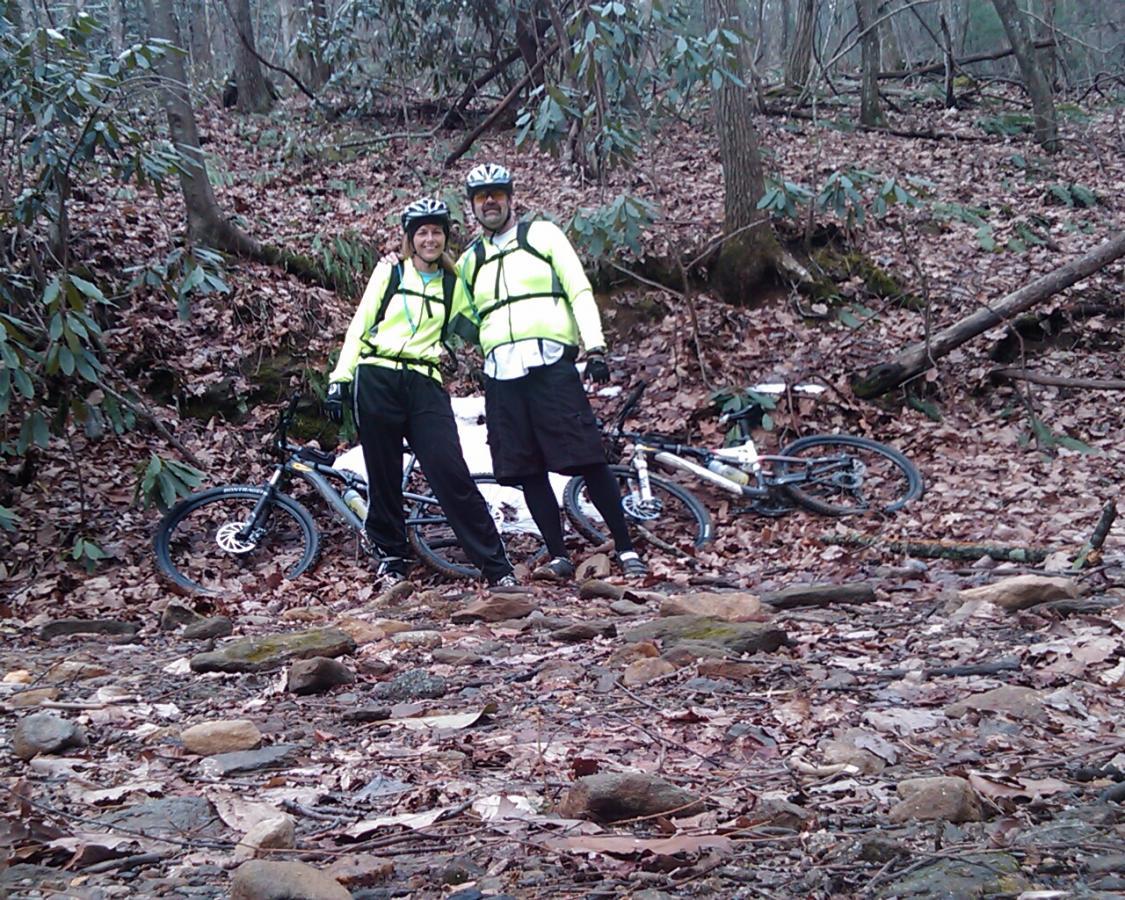 A man and woman wearing bright yellow cycling gear stand together in a wooded area covered with fallen leaves and rocks. Their mountain bikes are positioned nearby, partially obscured by foliage. The trail is rugged, indicating an adventurous biking excursion in nature. Bull / Jake Mountain mountain bike trail.