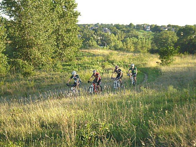 Four cyclists riding mountain bikes along a grassy trail in a scenic outdoor setting, with trees and rolling hills in the background. The sun is shining, creating a bright and lively atmosphere. Tranquility mountain bike trail.