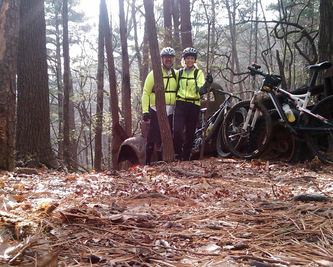 A pair of mountain bikers wearing bright yellow jackets and helmets, standing together in a wooded area. They are smiling beside their bicycles, with fallen leaves and pine needles covering the ground. In the background, there are trees and remnants of an old metal structure. Bull / Jake Mountain mountain bike trail.