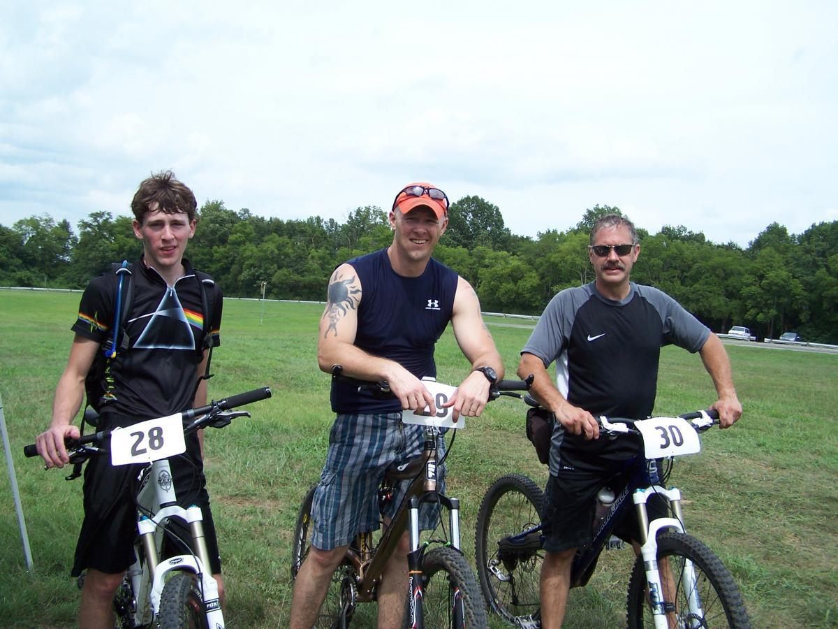 Three men stand with their mountain bikes in a grassy field on a cloudy day. The first man on the left wears a black shirt with a colorful design, holding his bike with the number 28 attached. The second man in the middle, wearing a sleeveless dark shirt and plaid shorts, has the number 29. The third man on the right, dressed in a gray and black athletic shirt, holds his bike with the number 30. Trees are visible in the background, and a road can be seen in the distance. Lock 4 mountain bike trail.