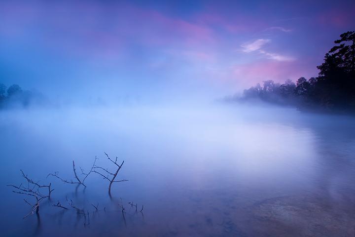 A serene landscape featuring a misty lake at dawn, with soft pastel colors in the sky and silhouettes of trees along the shore. In the foreground, a few bare branches extend partially into the water, creating a tranquil and ethereal atmosphere. Arrowhead Park mountain bike trail.