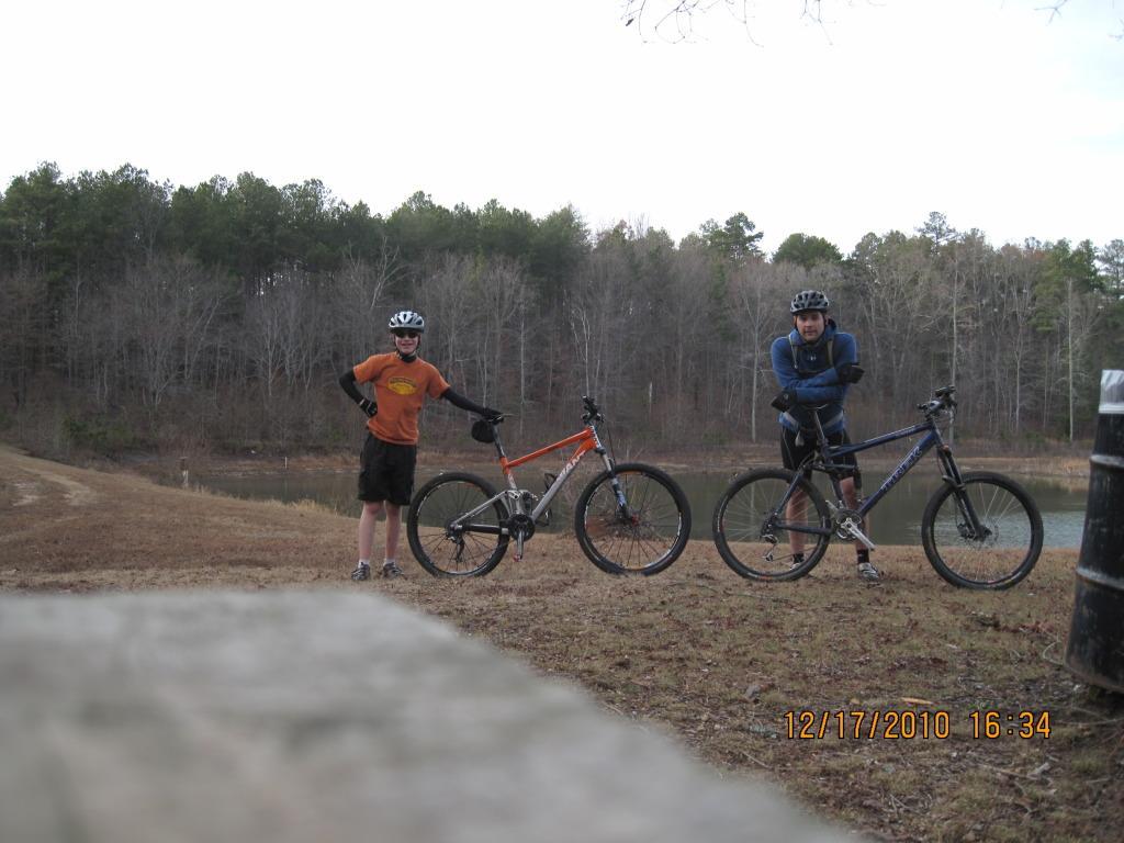 Two individuals stand next to their mountain bikes by a pond in a wooded area. One person, wearing an orange t-shirt and shorts, has their hands on their hips, while the other, dressed in a dark blue long-sleeve shirt and pants, crosses their arms. The background features trees with bare branches, indicating winter, and a cloudy sky above. Pilchers Pond mountain bike trail.