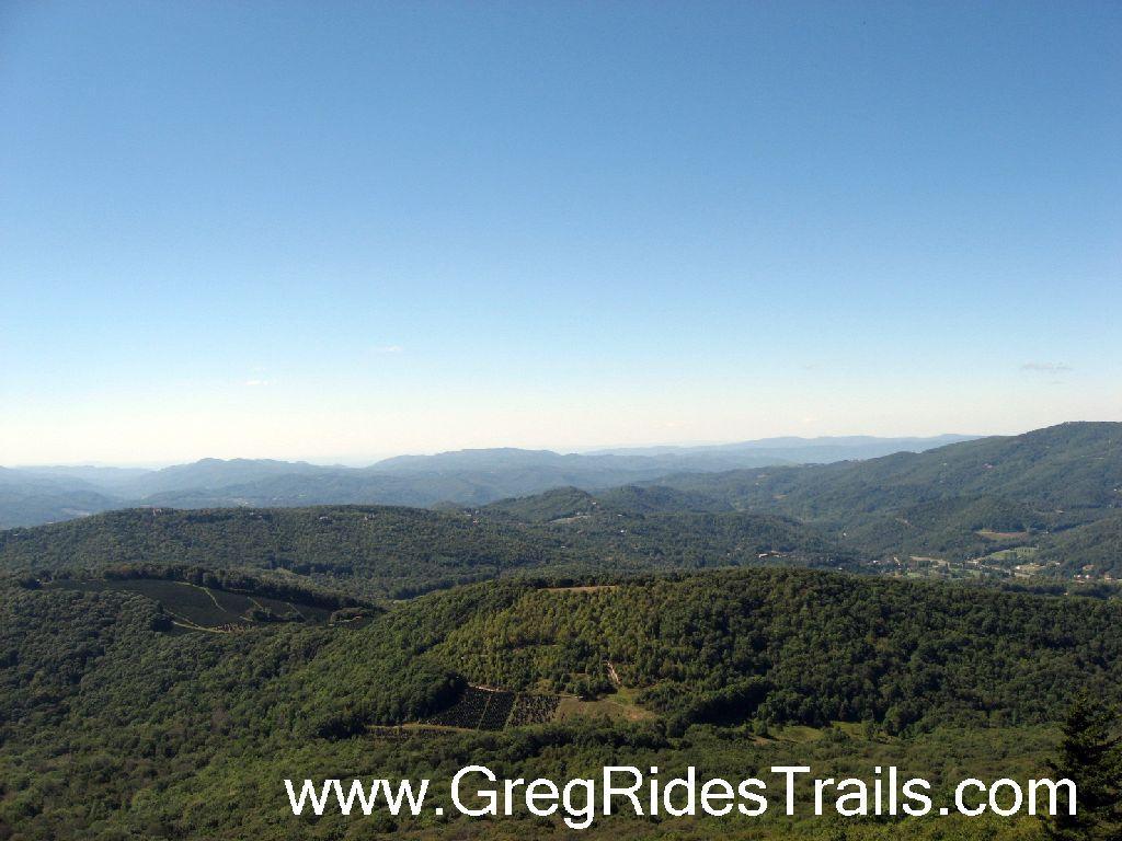A panoramic view of rolling green hills and distant mountains under a clear blue sky. The landscape features a mix of tree-covered slopes and open fields, creating a serene natural scene. Sugar Mountain Resort mountain bike trail.