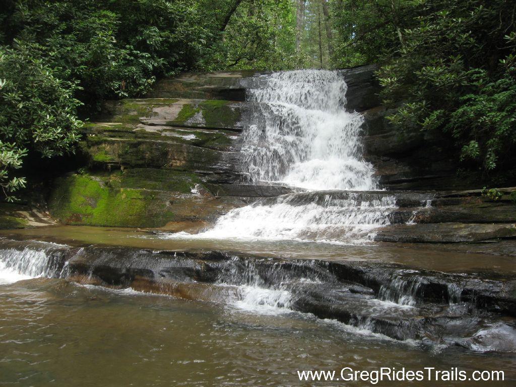 A beautiful waterfall cascading down moss-covered rocks into a serene pool, surrounded by lush green foliage and trees. Stonewall Falls Loop mountain bike trail.