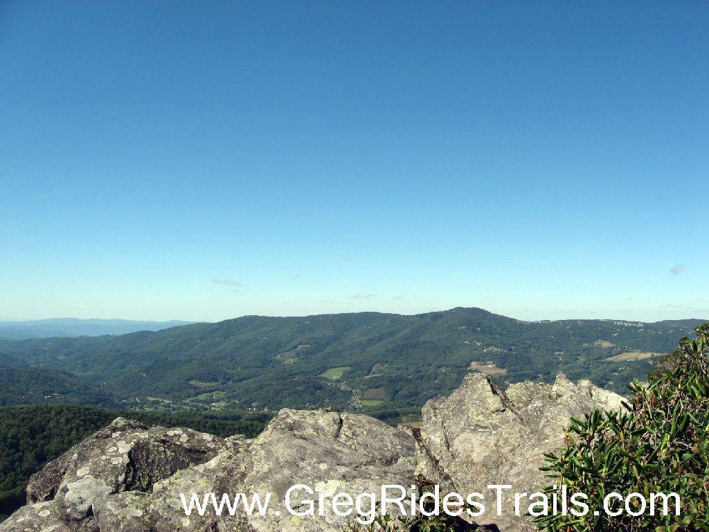 Panoramic view of rolling hills and valleys under a clear blue sky. Rocky foreground leads into lush green landscapes, showcasing the natural beauty of the mountains. Sugar Mountain Resort mountain bike trail.