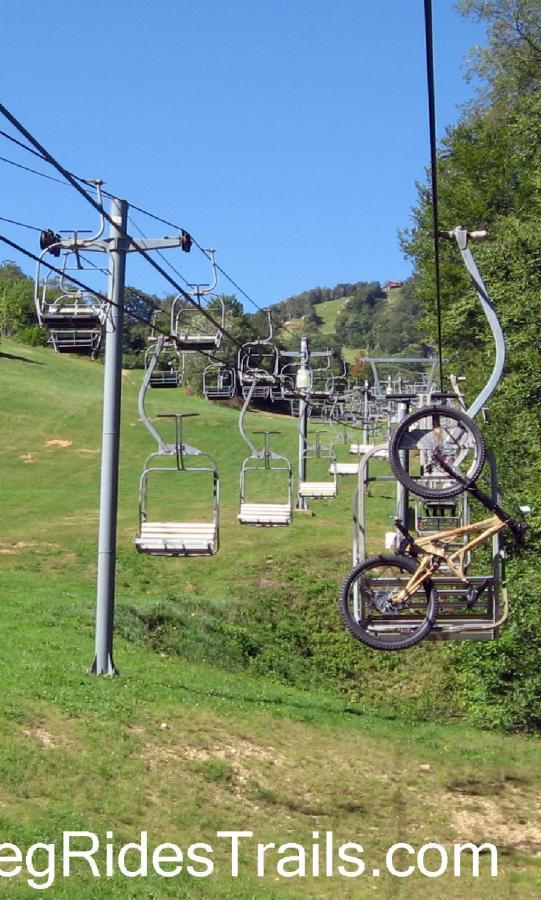 A series of empty chairlift seats on a sunny day, with a mountain bike hanging from one of the seats. The green hillside is visible in the background, showcasing a scenic outdoor environment. Sugar Mountain Resort mountain bike trail.