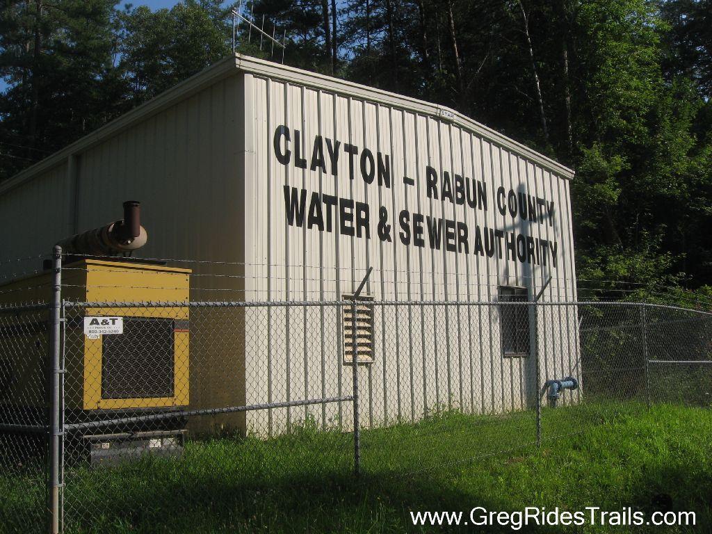 A metal building with the text "Clayton - Rabun County Water & Sewer Authority" prominently displayed on the side. A yellow generator is visible next to the building, which is surrounded by a chain-link fence and greenery. Stonewall Falls Loop mountain bike trail.