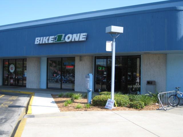 A storefront of a bicycle shop named "Bike 1 Lane," featuring large glass windows displaying bicycles inside. The building has a blue exterior with a prominent sign. A small parking lot is in front, along with some landscaping and a bicycle rack. The shop appears bright and inviting, indicating business hours and promotions.