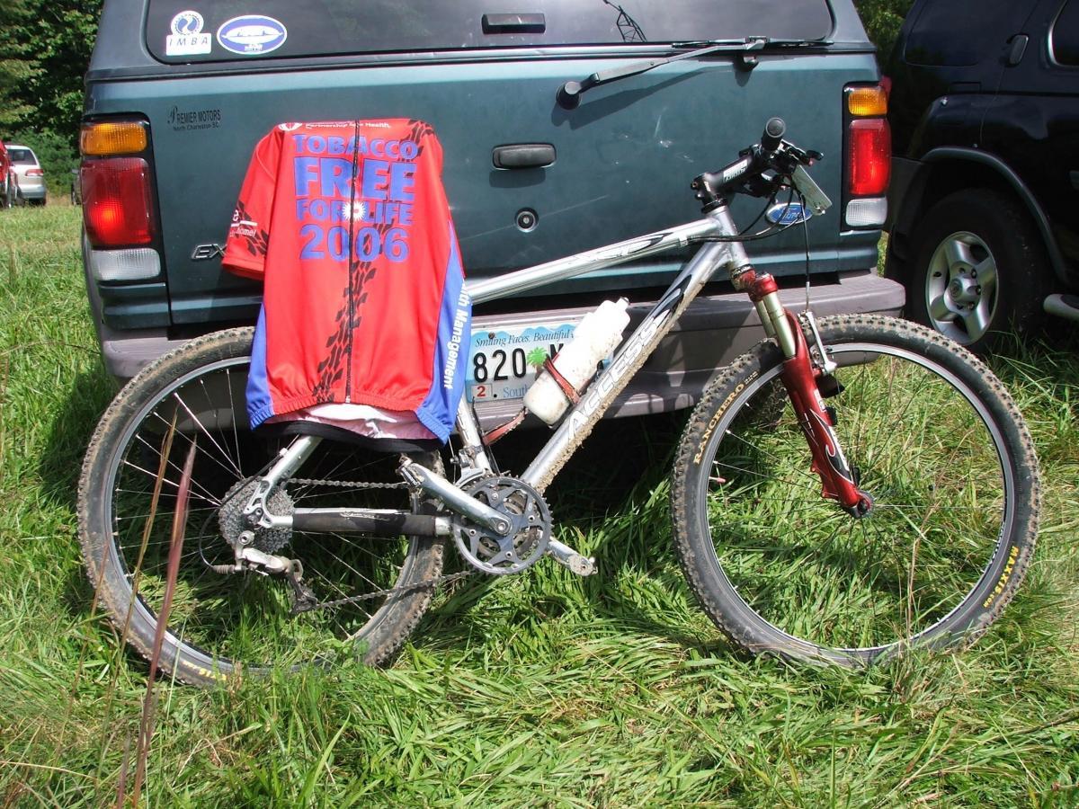 A mountain bike leaning against a parked vehicle in a grassy area, with a red and blue cycling jersey hanging on the bike. The jersey features the text "TOBACCO FREE FOR LIFE 2006." DuPont State Forest mountain bike trail.