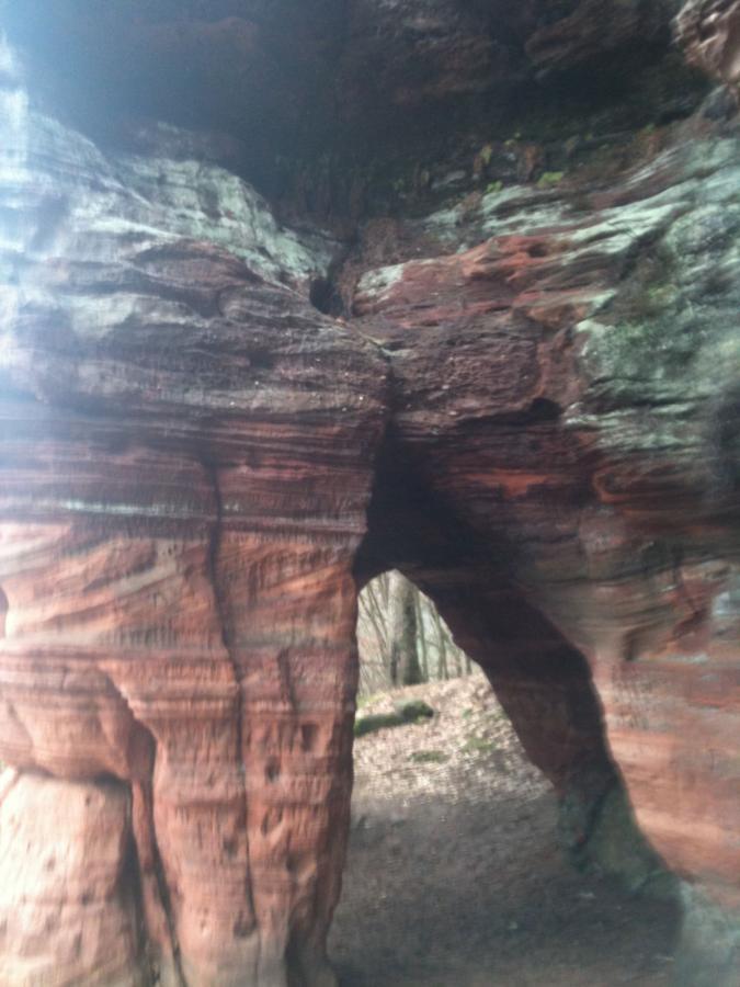 A natural rock formation with layered red and brown stone, featuring a large archway that leads into a wooded area. The image captures the texture and colors of the rock, with greenery in the background. Rodalben F Trail mountain bike trail.