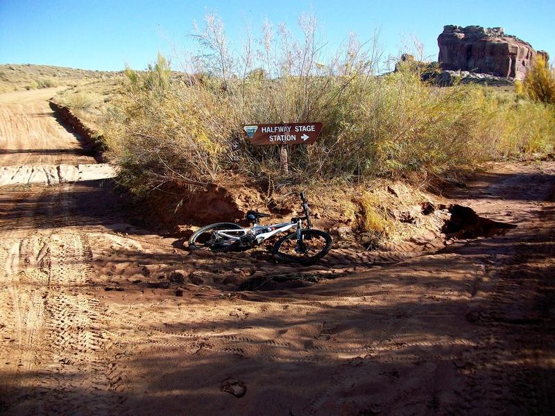 A mountain bike lies on its side next to a directional sign for "Halfway Stage Station." The scene features a dirt path lined with sparse vegetation in a rugged landscape, with rock formations visible in the background under a clear blue sky. Monitor &amp; Merrimac/Courthouse Pasture mountain bike trail.