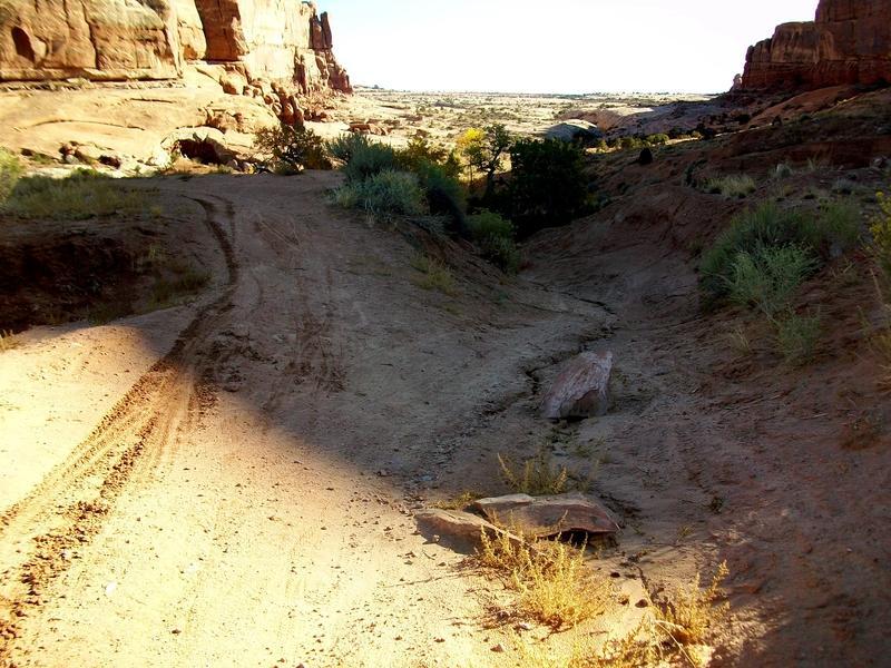 A dirt path winding through a rocky landscape with sparse vegetation, featuring tracks from vehicles in the foreground. In the background, tall rock formations and a vast open area under a clear sky. Monitor &amp; Merrimac/Courthouse Pasture mountain bike trail.