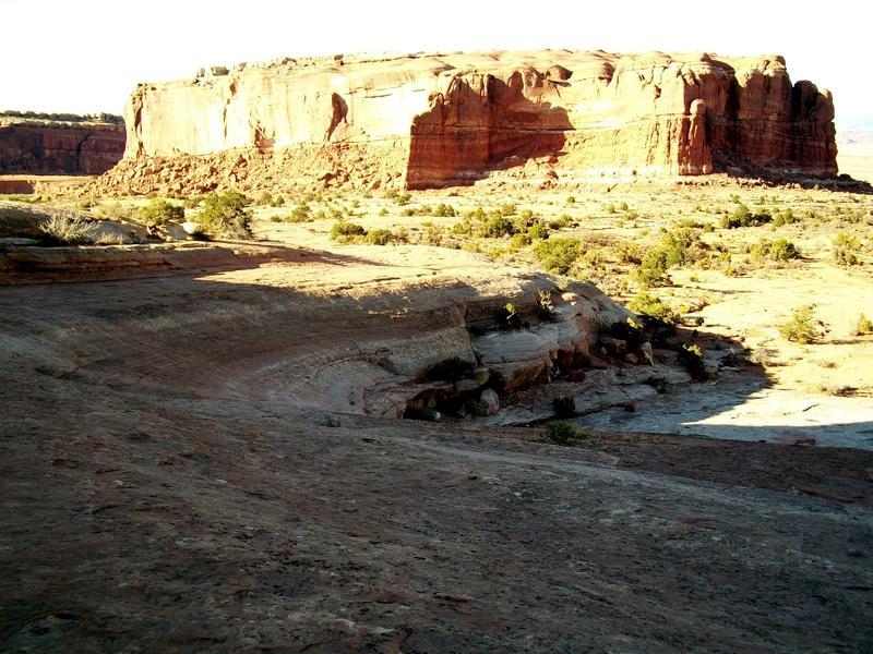 A panoramic view of a rocky landscape featuring a prominent red sandstone cliff, set against a clear blue sky. The foreground showcases rocky terrain with patches of vegetation, while the background highlights the towering cliff face illuminated by sunlight. Monitor &amp; Merrimac/Courthouse Pasture mountain bike trail.