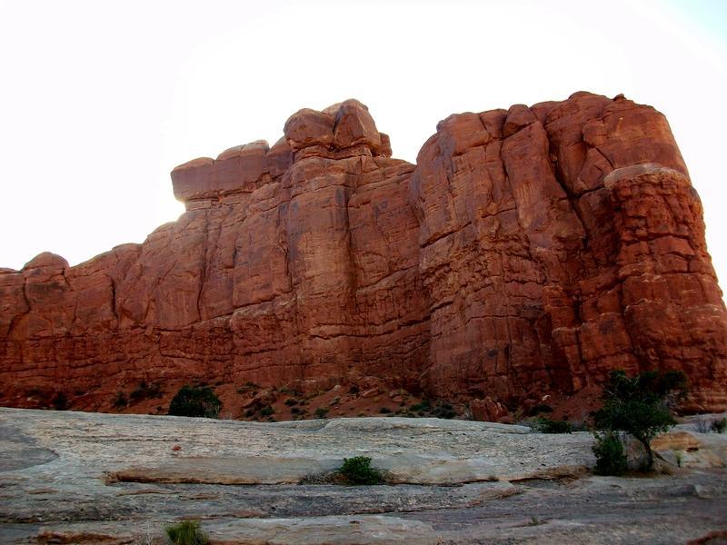 A large sandstone rock formation with steep, rugged cliffs and a warm, reddish hue, surrounded by sparse vegetation on a rocky terrain. The sky is bright in the background, highlighting the natural textures of the rock. Monitor &amp; Merrimac/Courthouse Pasture mountain bike trail.