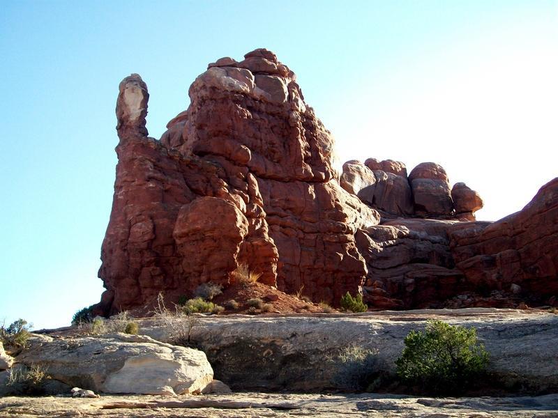Alt text: A towering red rock formation against a clear blue sky, with unique shapes and textures, surrounded by sparse vegetation and rocky terrain. Monitor &amp; Merrimac/Courthouse Pasture mountain bike trail.