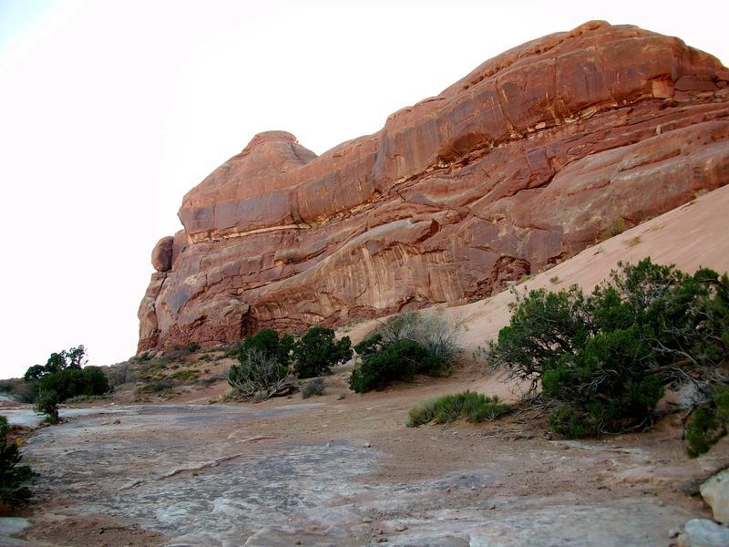A desert landscape featuring a large, rugged rock formation with layered red and brown textures. In the foreground, sparse vegetation including low shrubs and grasses contrasts with the rocky terrain. The sky above is bright, indicating daylight. Monitor &amp; Merrimac/Courthouse Pasture mountain bike trail.