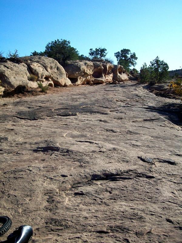 A rocky trail leading upward, surrounded by sparse vegetation and trees against a clear blue sky. The terrain is mostly flat with visible rock formations and a hint of a path. A piece of footwear can be seen in the lower left corner. Monitor &amp; Merrimac/Courthouse Pasture mountain bike trail.