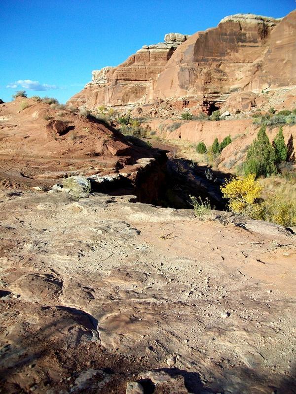 A rugged landscape featuring reddish rock formations and a deep canyon. The scene includes patches of greenery at the base of the cliffs, with a clear blue sky above, creating a striking contrast with the earthy tones of the terrain. Monitor &amp; Merrimac/Courthouse Pasture mountain bike trail.