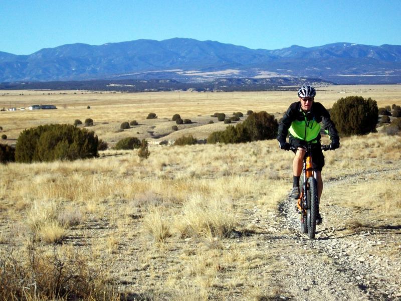 A person in a bright green and black outfit riding a mountain bike along a rocky trail in a grassy landscape, with mountains in the background under a clear blue sky. South Shore Lake Pueblo mountain bike trail.